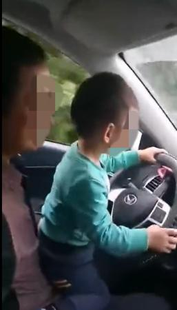 一男子让小孩手握汽车方向盘操作(图)